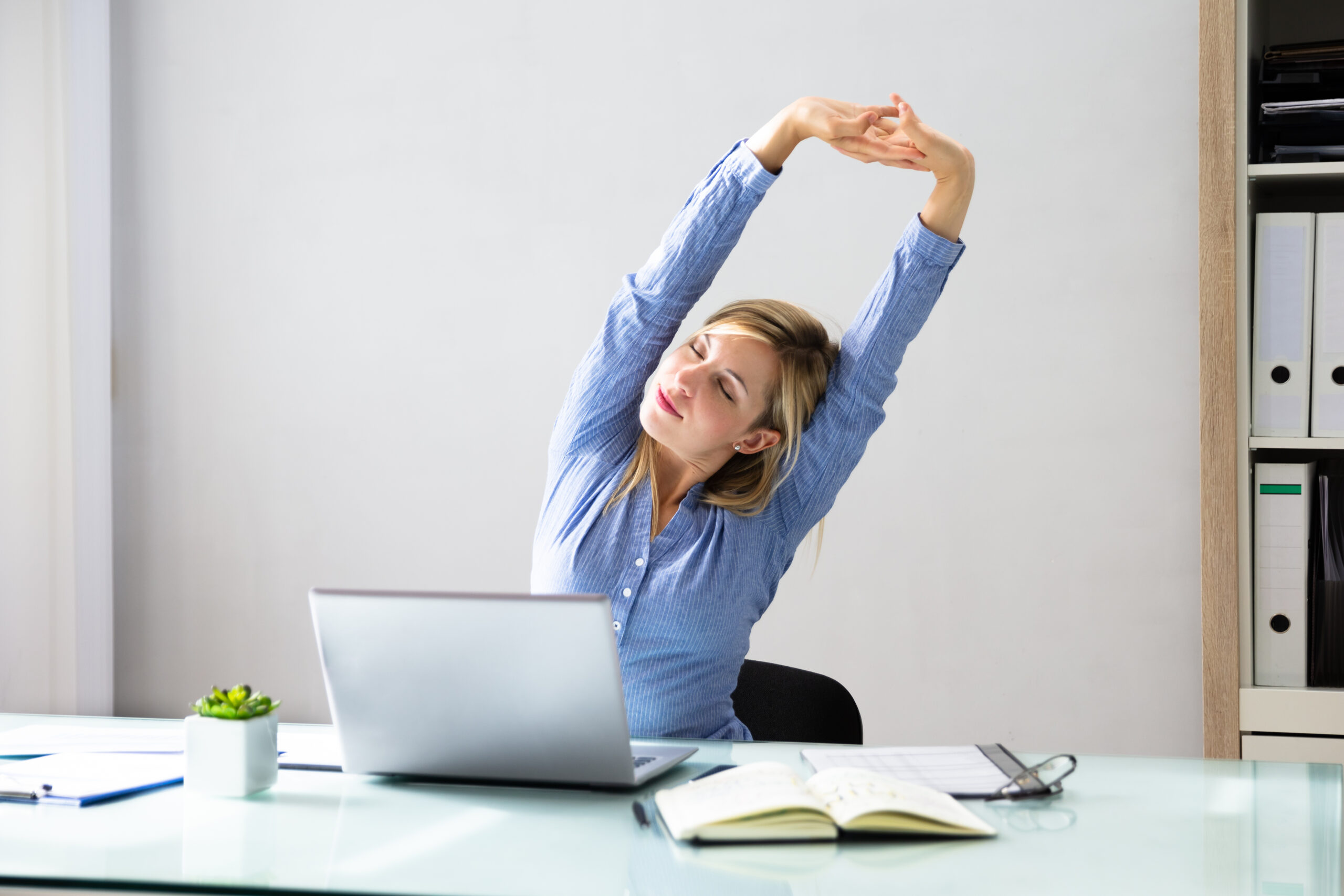 Foto zeigt eine Frau am Arbeitsplatz, die eine Dehnübung macht. 